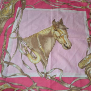 May include: A square silk scarf featuring a central illustration of a brown horse head wearing a bridle. The design includes tan leather equestrian tack, stirrups, and reins arranged in a border pattern against a vibrant pink background.