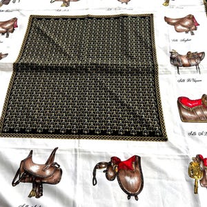 May include: A white silk scarf featuring a central black square panel with a repeating gold stirrup pattern. The border displays various brown leather horse saddles with red accents, each labeled with French text including Selle Arabe and Selle Anglaise.