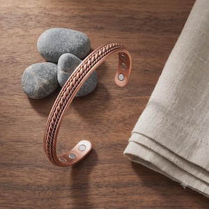 May include: A copper-toned metal cuff bracelet featuring a central twisted rope design. The interior of the open-ended bracelet displays several small, circular embedded magnets at both ends, providing a clear view of the construction.