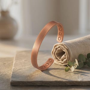 Può includere: Un bracciale a fascia in rame spazzolato con una finitura liscia e opaca. L'interno del bracciale presenta sei piccoli inserti magnetici circolari, con tre elementi posizionati su ciascuna delle estremità aperte della fascia metallica.