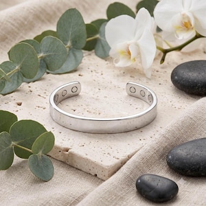 May include: A silver-toned metal cuff bracelet with a smooth, polished exterior surface. The interior of the open-ended band features several small, circular magnetic inserts embedded along both ends of the bracelet's inner circumference.