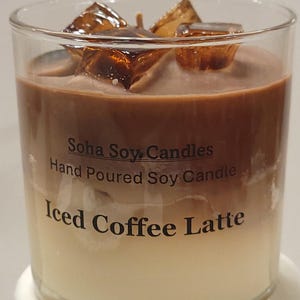 May include: A clear glass candle featuring a layered design resembling an iced coffee latte, with a dark brown top layer and a light cream bottom layer. The top is decorated with amber-colored wax ice cubes. Text reads Soha Soy Candles.
