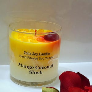 May include: A clear glass candle container filled with yellow and white wax, labeled Soha Soy Candles Hand Poured Soy Candle Mango Coconut Slush. The top features small yellow wax beads and a single dark red wax cherry with a red wick in the center.