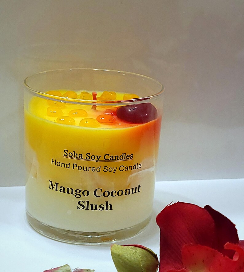 May include: A clear glass candle container filled with yellow and white wax, labeled Soha Soy Candles Hand Poured Soy Candle Mango Coconut Slush. The top features small yellow wax beads and a single dark red wax cherry with a red wick in the center.