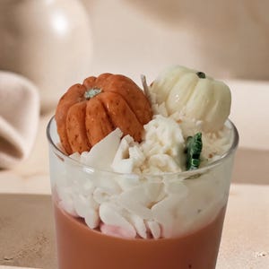 May include: A glass candle container filled with brown wax, topped with white wax shavings resembling whipped cream, a small orange pumpkin-shaped wax piece, a small white pumpkin-shaped wax piece, and a small green leaf-shaped wax accent.