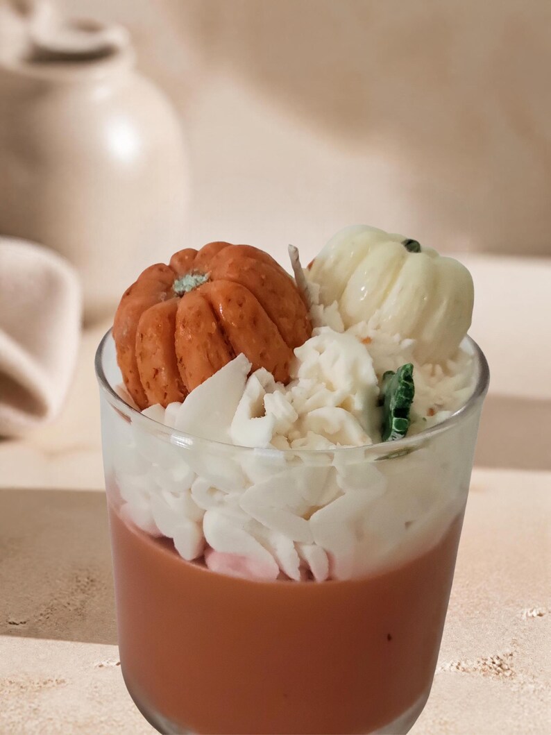May include: A glass candle container filled with brown wax, topped with white wax shavings resembling whipped cream, a small orange pumpkin-shaped wax piece, a small white pumpkin-shaped wax piece, and a small green leaf-shaped wax accent.