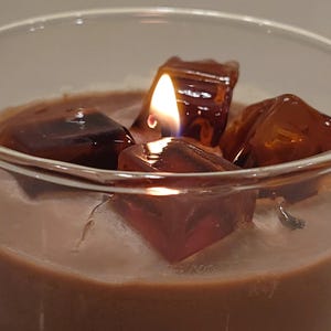 May include: A lit candle in a clear glass container featuring a brown wax base topped with several translucent, dark amber-colored wax cubes that resemble ice. The flame is visible between the wax cubes, casting a warm glow on the surface.