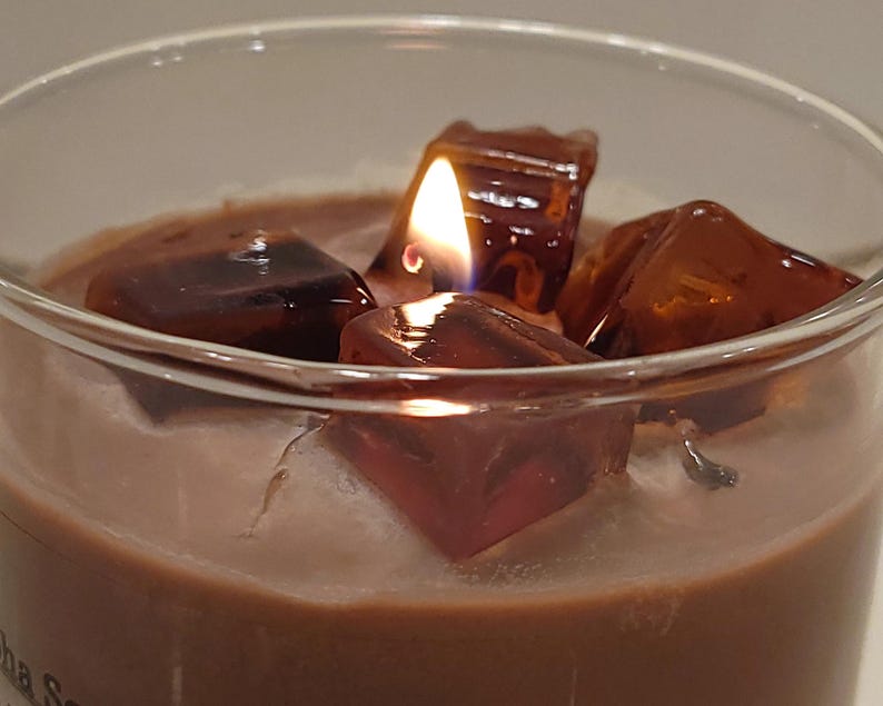 May include: A lit candle in a clear glass container featuring a brown wax base topped with several translucent, dark amber-colored wax cubes that resemble ice. The flame is visible between the wax cubes, casting a warm glow on the surface.