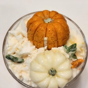 May include: A glass candle container filled with white wax, topped with a textured orange pumpkin, a smooth cream-colored pumpkin, and small green leaf-shaped wax accents. A single white wick protrudes from the center of the wax surface.