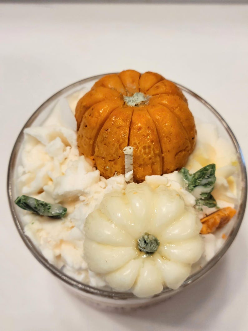 May include: A glass candle container filled with white wax, topped with a textured orange pumpkin, a smooth cream-colored pumpkin, and small green leaf-shaped wax accents. A single white wick protrudes from the center of the wax surface.