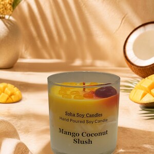May include: A clear glass jar candle with a layered yellow and white wax design. The label reads Soha Soy Candles Hand Poured Soy Candle Mango Coconut Slush. The top of the candle features small yellow wax droplets and a single dark red wax cherry.