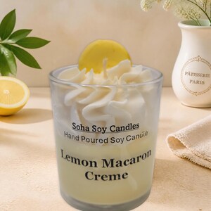 May include: A clear glass candle container filled with pale yellow soy wax, topped with white whipped wax swirls and a yellow circular wax accent. The label reads Soha Soy Candles Hand Poured Soy Candle Lemon Macaron Creme in black text.