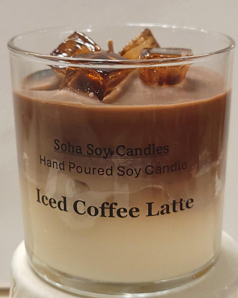 May include: A clear glass candle featuring a layered design resembling an iced coffee latte, with a dark brown top layer and a light cream bottom layer. The top is decorated with amber-colored wax ice cubes. Text reads Soha Soy Candles.