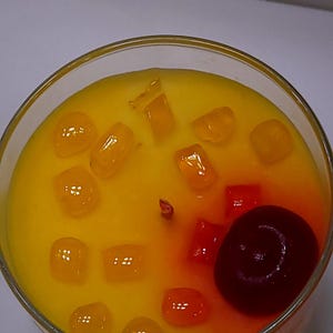 May include: A round glass container filled with yellow wax, featuring a central wick and topped with several small, translucent orange cubes and a single, dark red, round cherry-like wax accent positioned near the edge of the container.