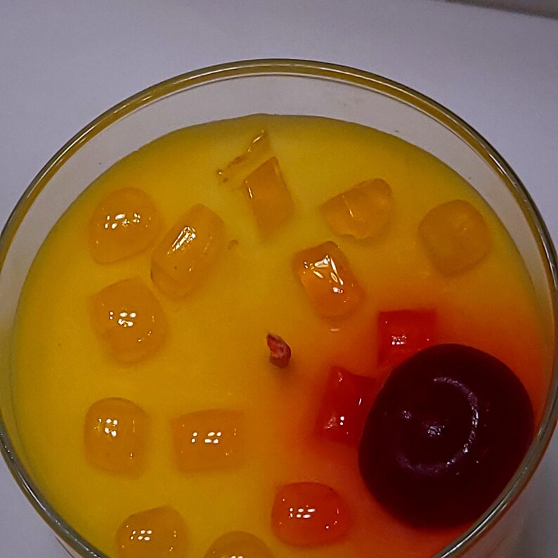 May include: A round glass container filled with yellow wax, featuring a central wick and topped with several small, translucent orange cubes and a single, dark red, round cherry-like wax accent positioned near the edge of the container.
