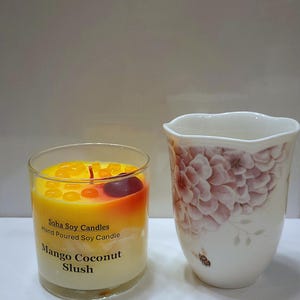May include: A clear glass candle container filled with yellow and white wax, topped with small orange wax droplets and a dark red wax cherry. The label reads Soha Soy Candles Hand Poured Soy Candle Mango Coconut Slush. Beside it is a white ceramic cup.