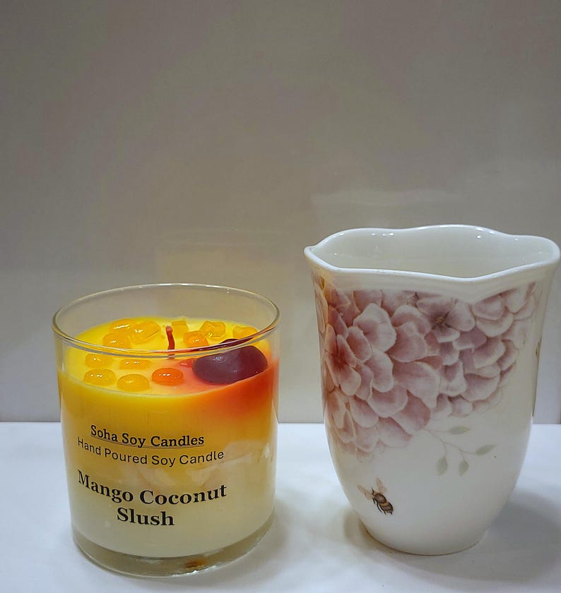 May include: A clear glass candle container filled with yellow and white wax, topped with small orange wax droplets and a dark red wax cherry. The label reads Soha Soy Candles Hand Poured Soy Candle Mango Coconut Slush. Beside it is a white ceramic cup.