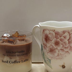 May include: A clear glass candle with a layered brown and cream wax design, topped with amber-colored wax cubes. The label reads Soha Soy Candles Hand Poured Soy Candle Iced Coffee Latte. Beside it is a white ceramic mug with pink hydrangea floral patterns.