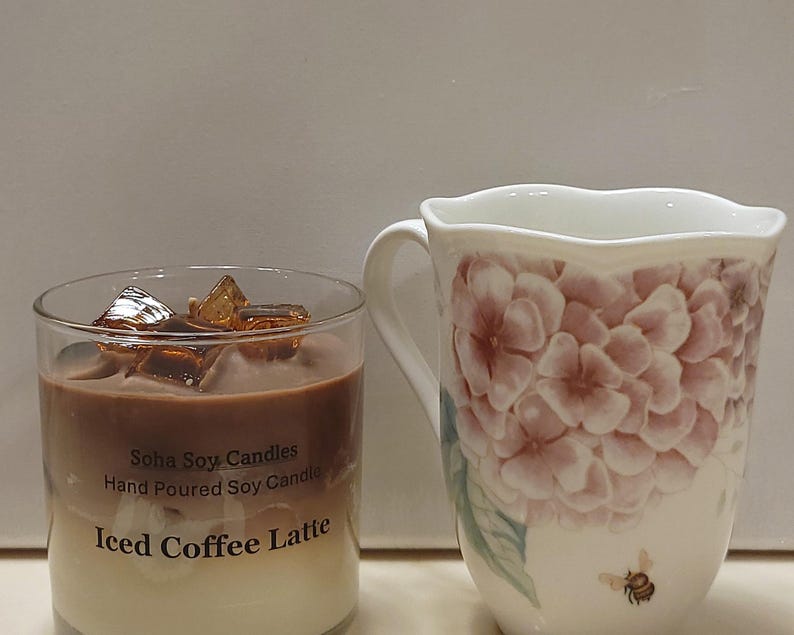 May include: A clear glass candle with a layered brown and cream wax design, topped with amber-colored wax cubes. The label reads Soha Soy Candles Hand Poured Soy Candle Iced Coffee Latte. Beside it is a white ceramic mug with pink hydrangea floral patterns.
