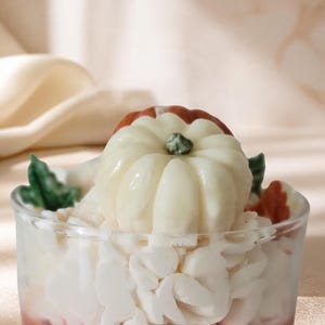 May include: A glass candle featuring a light brown base, topped with white wax piped to resemble whipped cream. A small, off-white pumpkin with a green stem sits in the center, accented by small green and orange wax leaves on the sides.
