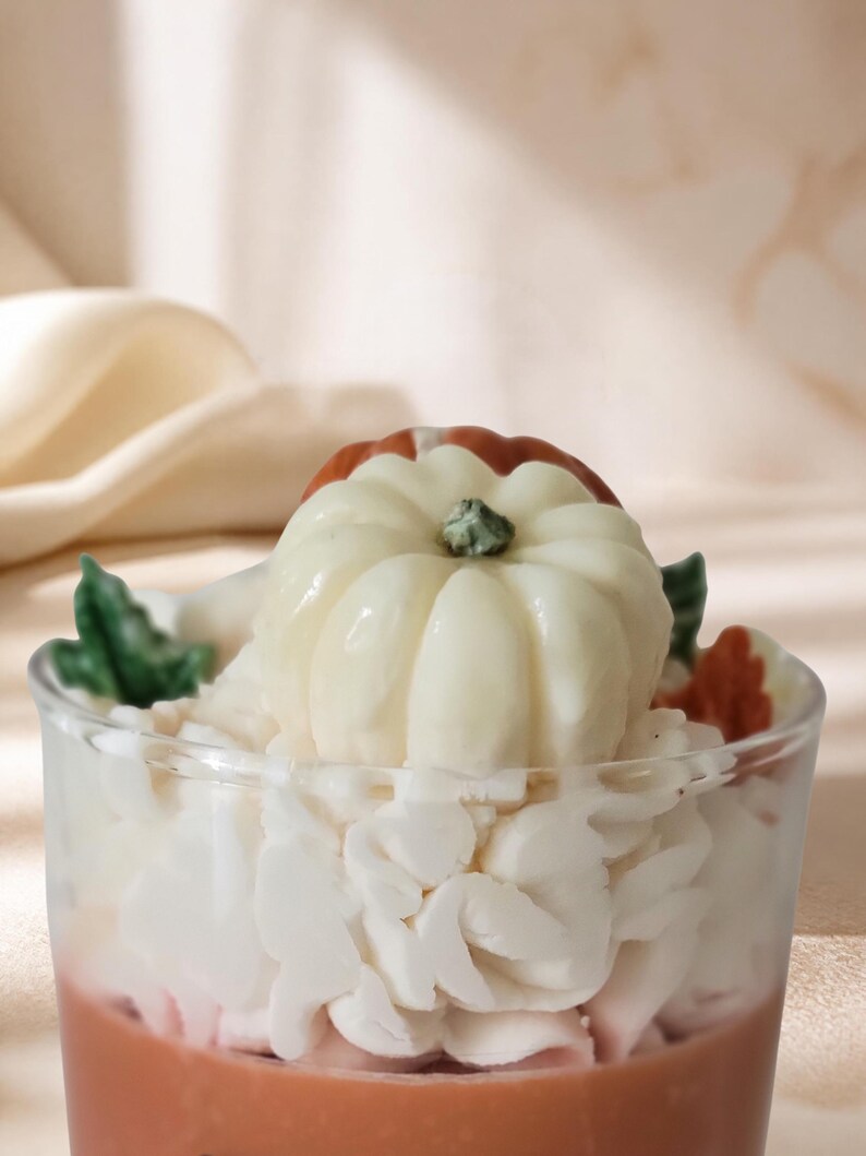 May include: A glass candle featuring a light brown base, topped with white wax piped to resemble whipped cream. A small, off-white pumpkin with a green stem sits in the center, accented by small green and orange wax leaves on the sides.
