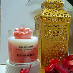 May include: A clear glass candle jar with a pink rose-shaped wax topper and a white and pink ombre wax base. The label reads Soha Soy Candles, Hand Poured Soy Candle, Princess Rose. A gold metal lantern with intricate cutouts stands behind it.