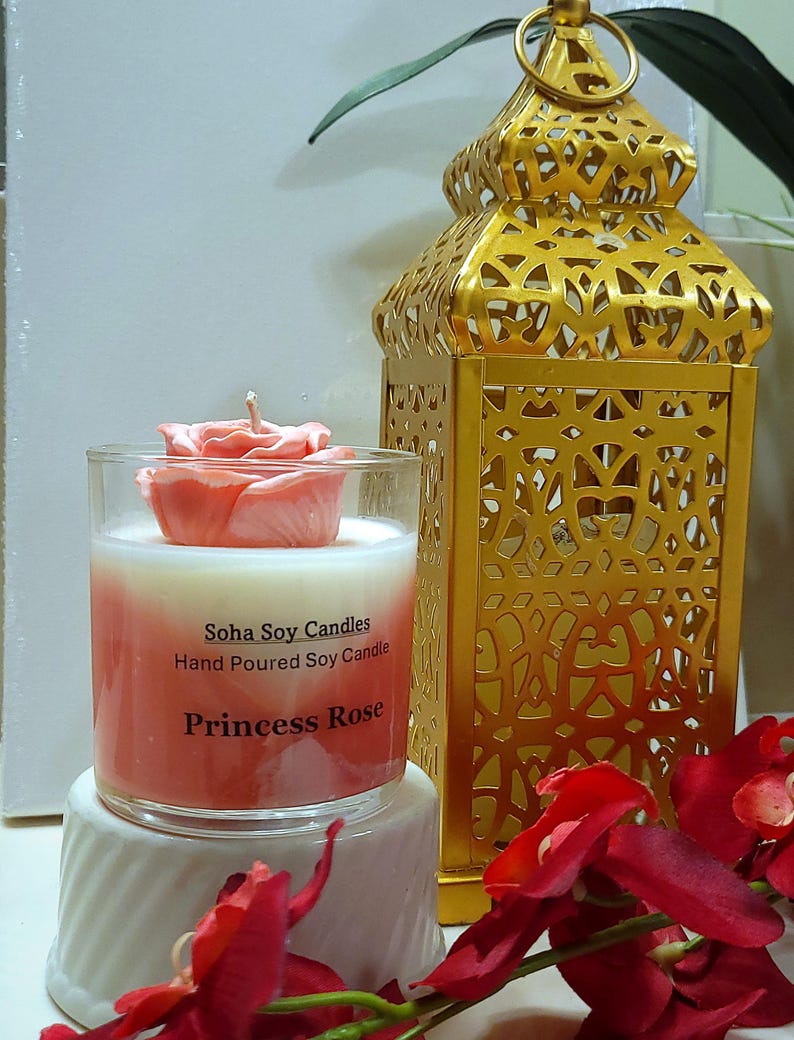 May include: A clear glass candle jar with a pink rose-shaped wax topper and a white and pink ombre wax base. The label reads Soha Soy Candles, Hand Poured Soy Candle, Princess Rose. A gold metal lantern with intricate cutouts stands behind it.