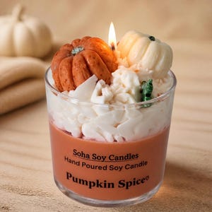 May include: A clear glass jar candle with orange wax, topped with white wax shavings and two small pumpkin-shaped wax embeds. The label reads: Soha Soy Candles, Hand Poured Soy Candle, Pumpkin Spice. A single lit wick is visible in the center.