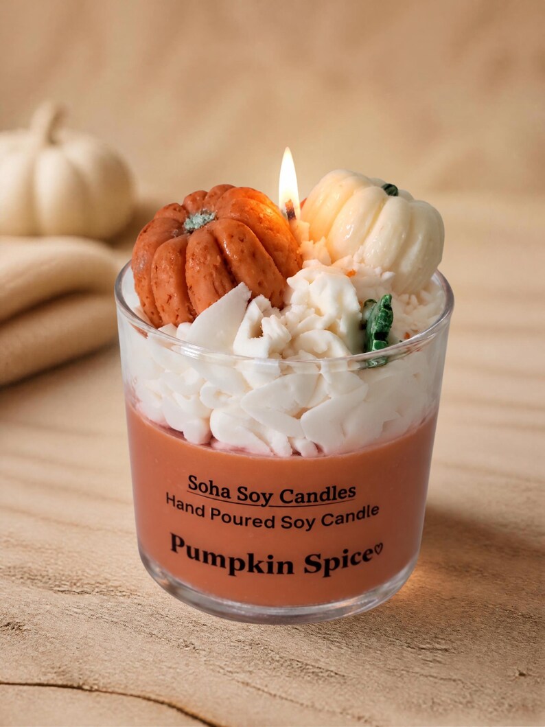 May include: A clear glass jar candle with orange wax, topped with white wax shavings and two small pumpkin-shaped wax embeds. The label reads: Soha Soy Candles, Hand Poured Soy Candle, Pumpkin Spice. A single lit wick is visible in the center.