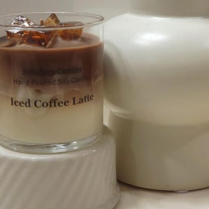 May include: A clear glass candle with layers of white and brown wax, topped with amber-colored wax cubes. The label reads Soho Soy Candles, Hand Poured Soy Candle, and Iced Coffee Latte. The candle sits on a white, ribbed, cylindrical base.