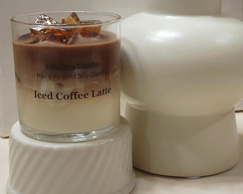 May include: A clear glass candle with layers of white and brown wax, topped with amber-colored wax cubes. The label reads Soho Soy Candles, Hand Poured Soy Candle, and Iced Coffee Latte. The candle sits on a white, ribbed, cylindrical base.