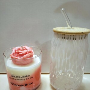 May include: A clear glass candle jar with a pink rose-shaped wax top, labeled Soha Soy Candles Hand Poured Soy Candle Princess Rose, next to a clear glass tumbler with a white speckled pattern, a bamboo lid, and a clear glass straw.