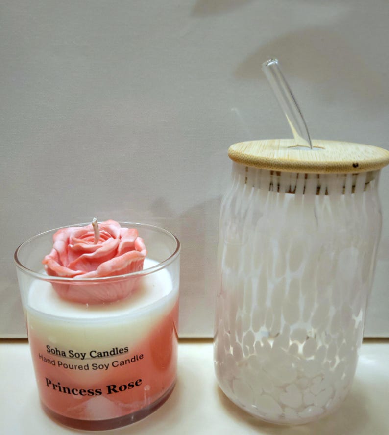 May include: A clear glass candle jar with a pink rose-shaped wax top, labeled Soha Soy Candles Hand Poured Soy Candle Princess Rose, next to a clear glass tumbler with a white speckled pattern, a bamboo lid, and a clear glass straw.