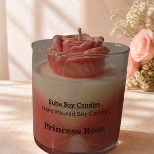 May include: A clear glass candle jar containing white soy wax with a pink rose-shaped wax topper. The label reads: Soha Soy Candles, Hand Poured Soy Candle, Princess Rose. The glass has a pink gradient tint, and a single cotton wick is visible.