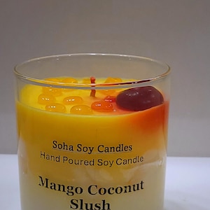 May include: A clear glass candle container filled with yellow and white wax. The label reads Soha Soy Candles, Hand Poured Soy Candle, Mango Coconut Slush. The top features small yellow and orange wax droplets and a single dark red wax piece.