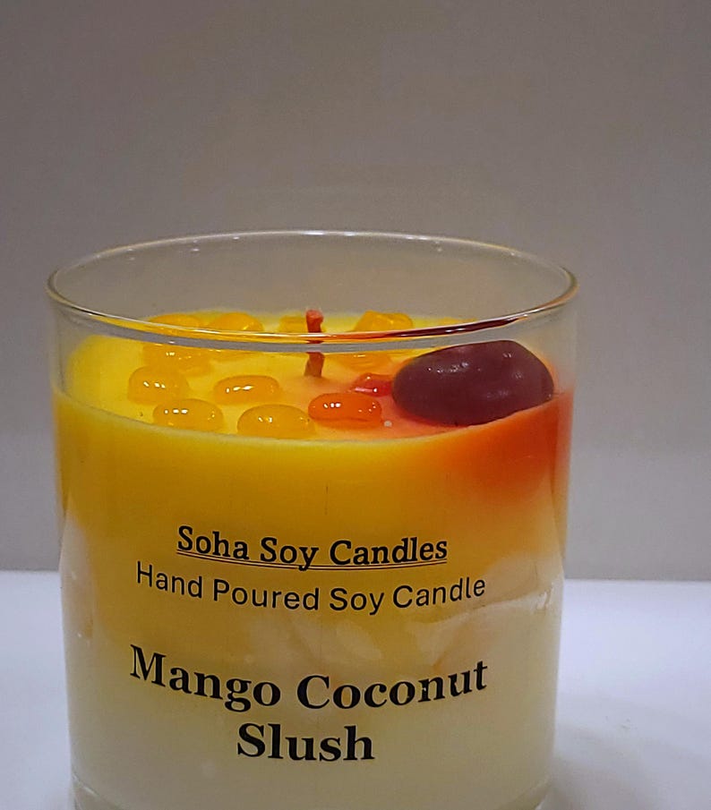 May include: A clear glass candle container filled with yellow and white wax. The label reads Soha Soy Candles, Hand Poured Soy Candle, Mango Coconut Slush. The top features small yellow and orange wax droplets and a single dark red wax piece.