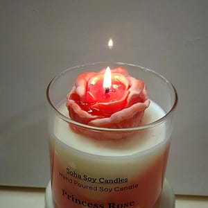 May include: A clear glass jar candle with a pink rose-shaped wax topper and a lit wick. The label reads Soha Soy Candles, Hand Poured Soy Candle, and Princess Rose. The wax inside the jar is white, transitioning to a soft pink hue at the base.