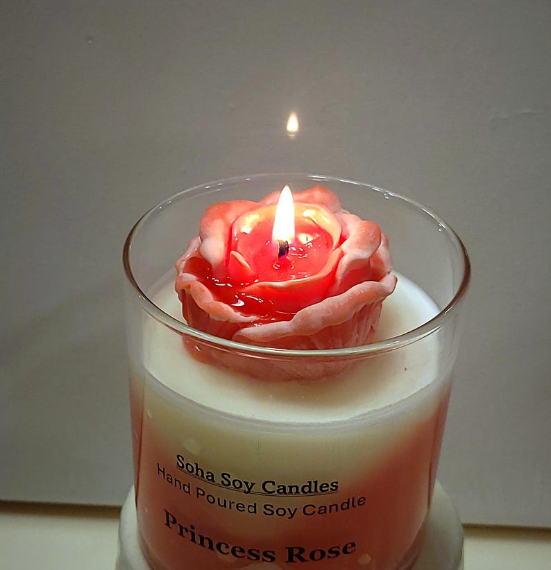 May include: A clear glass jar candle with a pink rose-shaped wax topper and a lit wick. The label reads Soha Soy Candles, Hand Poured Soy Candle, and Princess Rose. The wax inside the jar is white, transitioning to a soft pink hue at the base.