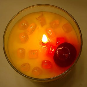May include: A round glass candle container filled with yellow wax, featuring a single lit wick in the center. The surface is decorated with several small, translucent orange and red wax cubes, along with one large, smooth, dark red wax sphere.