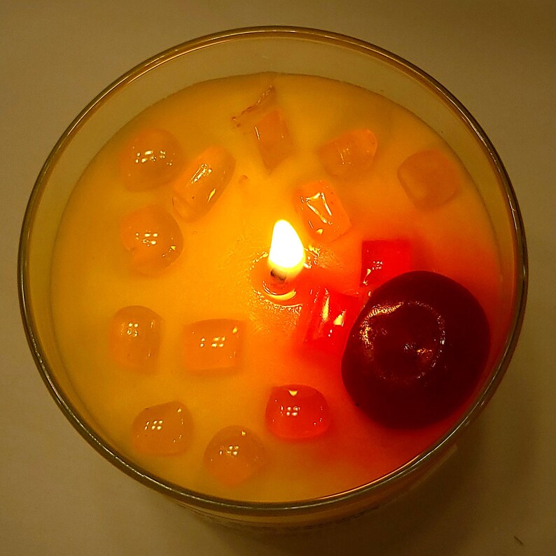 May include: A round glass candle container filled with yellow wax, featuring a single lit wick in the center. The surface is decorated with several small, translucent orange and red wax cubes, along with one large, smooth, dark red wax sphere.