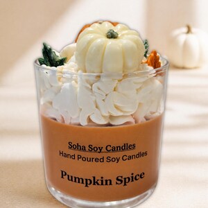 May include: A clear glass candle filled with orange wax, topped with white whipped wax and a small white pumpkin decoration. The label reads Soha Soy Candles, Hand Poured Soy Candles, and Pumpkin Spice in black text on the front of the glass.