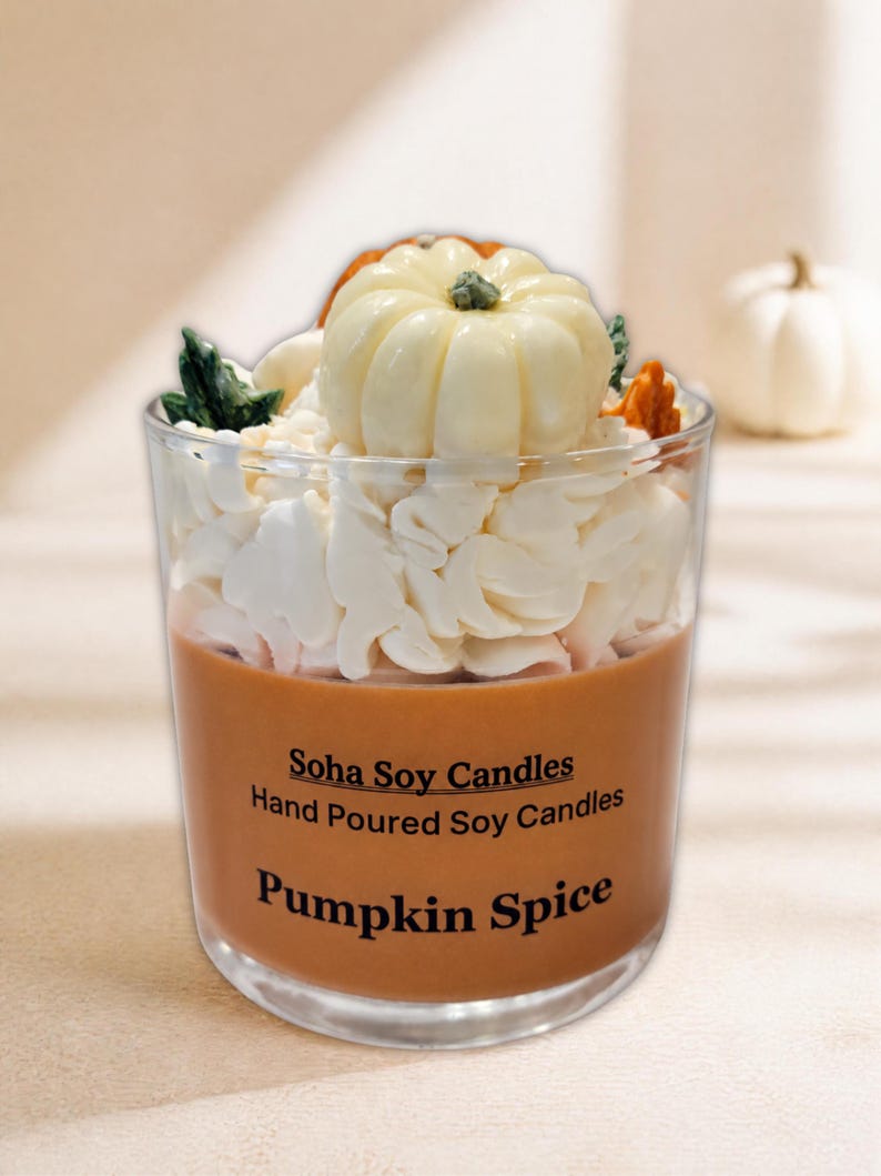 May include: A clear glass candle filled with orange wax, topped with white whipped wax and a small white pumpkin decoration. The label reads Soha Soy Candles, Hand Poured Soy Candles, and Pumpkin Spice in black text on the front of the glass.