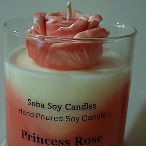 May include: A clear glass candle container filled with white and coral-colored wax, topped with a coral-colored rose-shaped wax ornament. The label reads: Soha Soy Candles, Hand Poured Soy Candle, Princess Rose. A single cotton wick protrudes.
