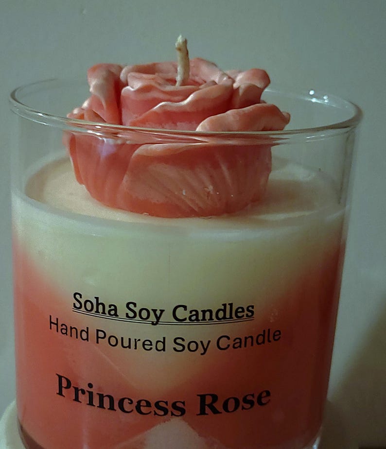 May include: A clear glass candle container filled with white and coral-colored wax, topped with a coral-colored rose-shaped wax ornament. The label reads: Soha Soy Candles, Hand Poured Soy Candle, Princess Rose. A single cotton wick protrudes.