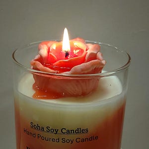 May include: A clear glass jar candle with a white wax base and an orange gradient. A peach-colored rose-shaped wax topper sits on top, with a lit wick in the center. The label reads Soha Soy Candles Hand Poured Soy Candle with partial text below.