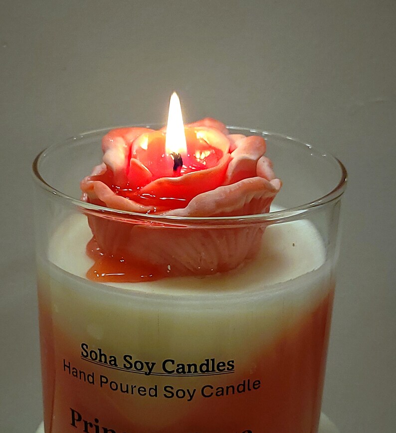 May include: A clear glass jar candle with a white wax base and an orange gradient. A peach-colored rose-shaped wax topper sits on top, with a lit wick in the center. The label reads Soha Soy Candles Hand Poured Soy Candle with partial text below.