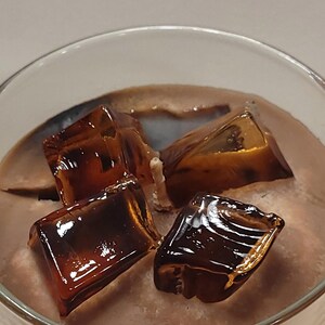 May include: A glass container filled with brown wax and topped with three translucent, dark amber-colored wax cubes that resemble ice. A single white cotton wick is positioned in the center of the wax, surrounded by the decorative wax ice cubes.