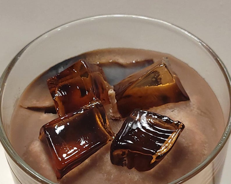 May include: A glass container filled with brown wax and topped with three translucent, dark amber-colored wax cubes that resemble ice. A single white cotton wick is positioned in the center of the wax, surrounded by the decorative wax ice cubes.