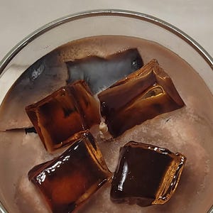 May include: A glass container filled with a light brown, textured substance resembling a beverage, topped with four dark brown, translucent, square-shaped wax pieces that look like ice cubes, and a single central wick protruding from the surface.