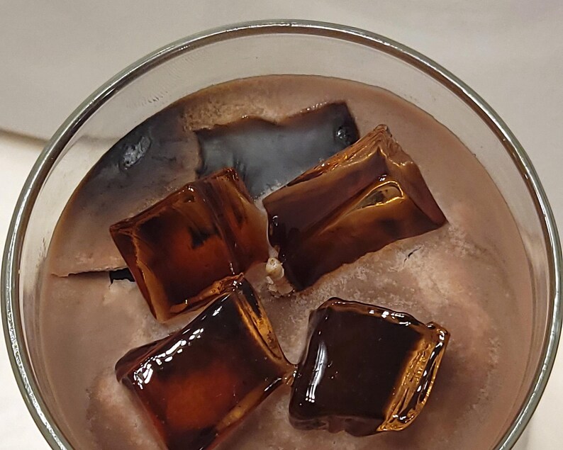 May include: A glass container filled with a light brown, textured substance resembling a beverage, topped with four dark brown, translucent, square-shaped wax pieces that look like ice cubes, and a single central wick protruding from the surface.